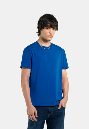 Uomo con capelli scuri che indossa una camicia blu a maniche corte, una collana a catena d'argento e jeans scuri, in piedi davanti a uno sfondo chiaro e uniforme.