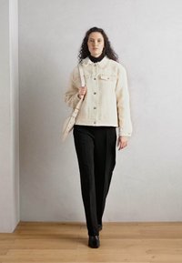Off-white Schafslederjacke mit Knopfverschluss und Taschen, kombiniert mit schwarzen Hosen und Stiefeln, vor einem neutralen Hintergrund.