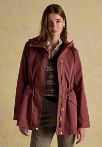 Bordowa wodoodporna kurtka z kapturem, z regulowanym pasem, złotymi detalami oraz dwoma bocznymi kieszeniami. Nosi ją osoba, która ma na sobie wzorzysty sweter i w kratkę spodnie.