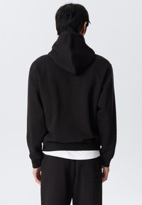 Persona vistiendo una sudadera negra y pantalones negros, de pie de espaldas contra un fondo gris claro liso.