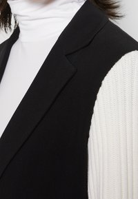 Blazer noir sans manches porté sur un pull en maille côtelée blanc et un haut blanc à col montant, mettant en valeur les détails du décolleté et des épaules.