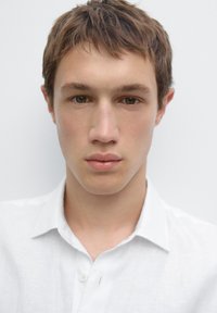 Porträt eines jungen Mannes mit kurzem braunem Haar, der ein weißes Hemd mit Kragen trägt. Er hat einen neutralen Gesichtsausdruck und braune Augen vor einem weißen Hintergrund.