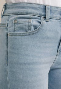 Helle blaue Denim-Jeans mit einer mittelhohen Taille, Fronttaschen und kontrastierendem gelben Stich, die eine glatte Textur und eine taillierte Passform zeigen.