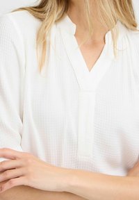 Hvid tekstureret bluse med V-udskæring og korte ærmer. Stoffet har et subtilt ternet mønster.