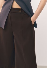 Pantalones a medida de color marrón con una textura suave, que cuentan con bolsillos laterales, cierre de botón y un corte relajado, combinados con una blusa azul oscuro.