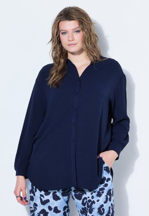 Femme aux cheveux blonds ondulés, portant une chemise ample bleu marine à boutons et un pantalon bleu clair avec des taches abstraites noires, debout contre un mur blanc.