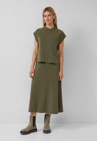 Kobieta ubrana w oliwkową bezrękawnik i pasującą długą spódnicę, w taupe ankle boots, stoi na tle jasnym.