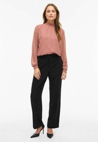 Bluzka w jasno różowym kolorze z długim rękawem, z fakturowanym wzorem jodełki i marszczonym dekoltem, zestawiona z czarnymi, dopasowanymi spodniami i czarnymi szpilkami o ostrym nosku.