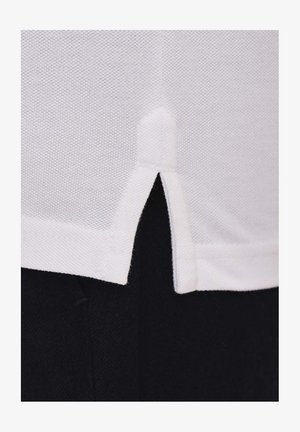 Biała bawełniana koszula o strukturalnym wykończeniu, z rozcięciem na dolnym brzegu. Tło jest ciemne, co kontrastuje z koszulą.