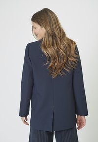 Blazer elegant de culoare bleumarin, cu mâneci lungi, croială dreaptă și ventilație la spate. Material fin cu un luciu subtil, asortat cu pantaloni cu dungi.