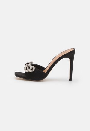 Mule à talon haut en daim noir avec bout ouvert et détail décoratif de chaîne argentée tressée sur la bride avant.