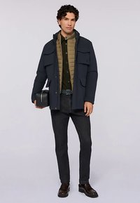 Giacca blu scuro con molteplici tasche frontali, sovrapposta a un gilet trapuntato marrone chiaro. Jeans neri e scarpe in pelle marrone completano l'outfit.