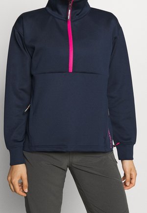 Pull-over bleu marine avec une demi-fermeture éclair rose et des poignets assortis, porté avec un pantalon gris foncé, montrant le haut du corps et une partie des mains.