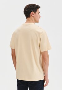 Beżowa bawełniana koszulka z okrągłym dekoltem i krótkimi rękawami. Z tyłu, w pobliżu dekoltu, znajduje się małe logo nadrukowane. Miękka tekstura.