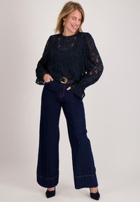 Marineblauwe kanten blouse met lange mouwen en gegolfde manchetten, gecombineerd met high-waisted wijde denim jeans met zichtbare stiksels en een riem.