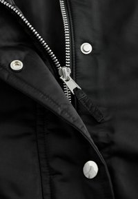 Schwarzer Stoff mit teilweise geöffnetem silbernem Reißverschluss und Metall-Druckknöpfen an einer Jacke oder einem Mantel.