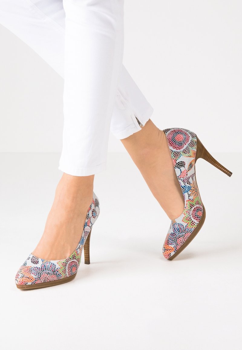 Jambes en pantalon blanc portant des chaussures à talons hauts à motifs mosaïque colorés avec des talons en bois stilettos sur un sol blanc.