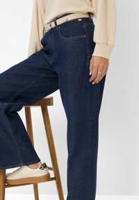 Donkerblauwe high-waisted jeans met een relaxte fit, voorzien van stiksels en een beige riem. Gecombineerd met witte sneakers en een houten kruk.