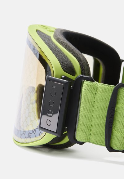 Ochelari de schi Gucci cu un cadru verde neon, lentilă transparentă cu un model logo și accente texturate negre pe curea și umplutură.