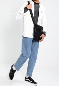 Svart axelväska i tyg med framficka, justerbar rem och jämn yta, i kombination med en vit skjorta och blå jeans.