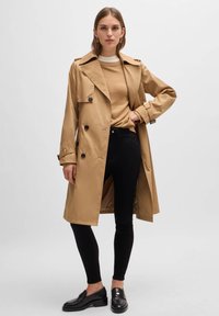 Beige Trenchcoat, doppelt knöpfbares Design, dunkle Knöpfe, breiter Kragen, taillierter Gurt, kombiniert mit schwarzen Hosen und Schuhen, minimalistischer Stil.