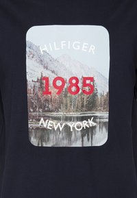 Námořnická modrá tričko má obdélníkovou grafiku s fotografií krajiny a textem "HILFIGER 1985 NEW YORK" v červené a bílé barvě.