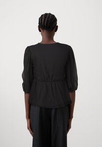 Svart, lättviktig topp med rynkad midja, genomskinliga puffärmar och rund halsringning, visad bakifrån. Slät textur, minimalistisk design.