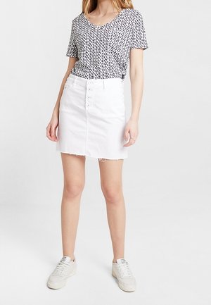 Donna che indossa un top a maniche corte con fantasia, una gonne in denim bianca con bottoni e sneaker bianche, in piedi su uno sfondo bianco.