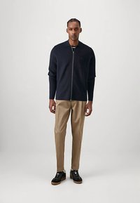 Mornarsko plava pletena jakna na patentni zatvarač s ovratnikom, uparena s bež hlačama i crnim tenisicama. Ima džep na prsima i teksturiranu tkaninu.