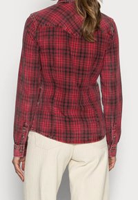 Camisa a cuadros rojos y negros con botones y mangas largas. Presenta un dobladillo curvado y puños con detalles de botones. Se lleva sobre pantalones beige de pierna ancha.