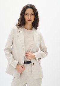 Beige linnblazer med enknappsspänning, spettslag och två framfickor. Kombinerad med en ljus topp och svart bälte.