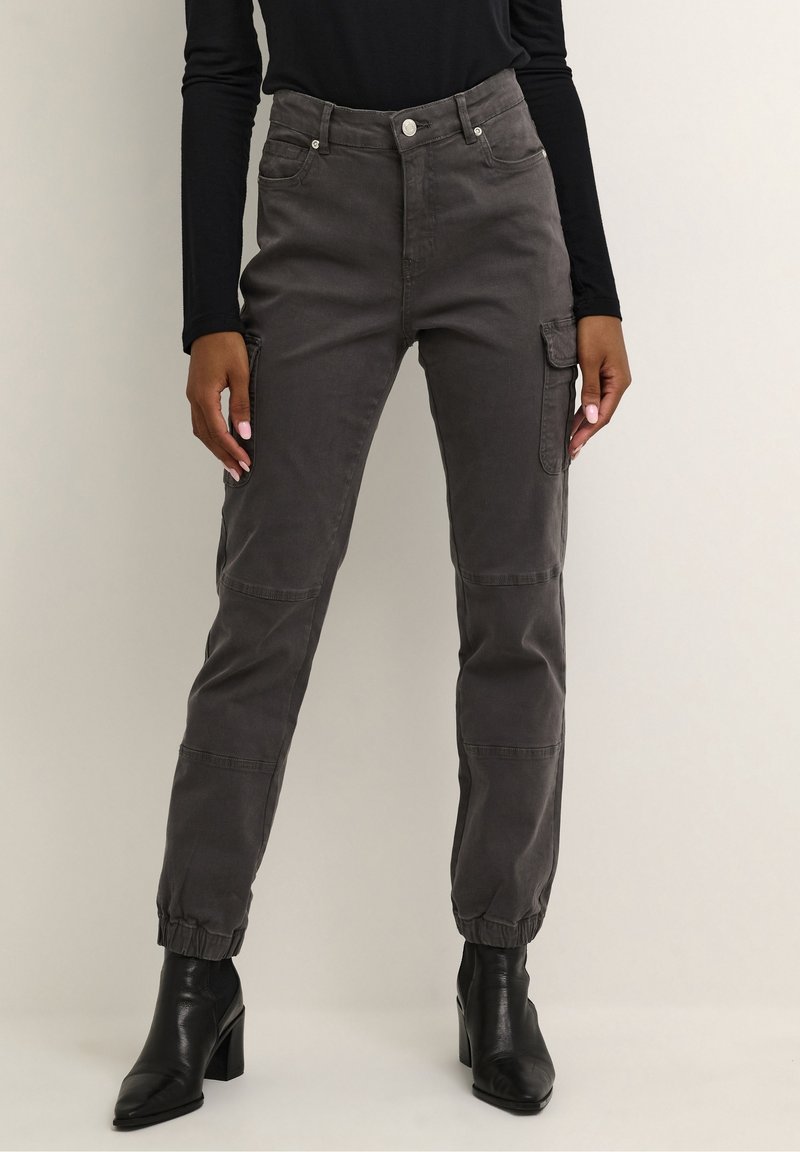Femme portant un pantalon cargo gris charbon avec des poignets élastiques et des bottines noires à talons, debout devant un fond uni.