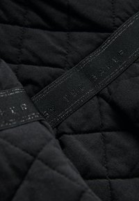 Černá prošívaná látka s lesklou černou štítkem s raženým textem "BY TED BAKER." Má měkkou texturu a vzor ve tvaru diamantů.