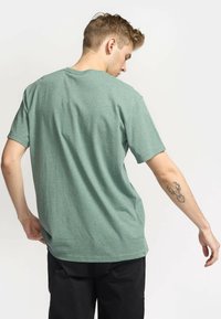 Groen gemêleerd katoenen T-shirt met een ronde hals, korte mouwen en een rechte snit. Opvallende tatoeage zichtbaar op de arm. Draagt zwarte broek.
