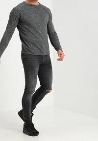 Mężczyzna w szaro-olikowym, długim rękawie koszuli, w podartych czarnych skinny jeansach i czarnych trampkach, stojący na tle jasnego tła.