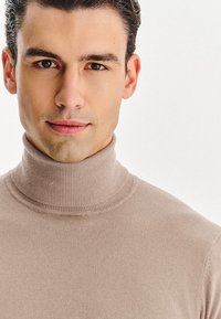 Beżowy sweter z golfem charakteryzuje się prążkowanym kołnierzem i gładką fakturą, ściśle dopasowany wokół szyi i klatki piersiowej, o prostym wzorze.