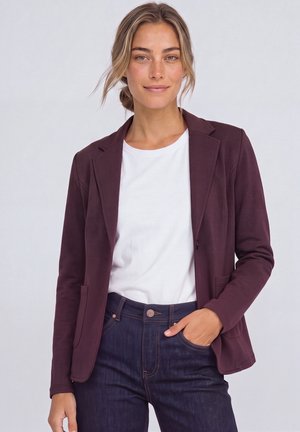 Femme aux cheveux châtain clair portant un blazer bordeaux, une chemise blanche et un jean bleu foncé, debout avec une main dans la poche devant un fond uni.