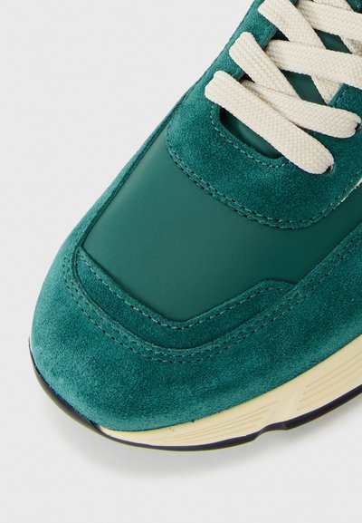Close-up of teal suede and leather sneaker toe with off-white laces and beige rubber sole on white background.