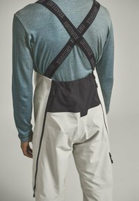 Weiße Latzhosen mit schwarzen Akzenten, verstellbaren Trägern und dem Markennamen "ORTOVOX", kombiniert mit einem strukturierten, türkisen Langarmshirt.