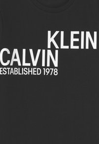 Černé bavlněné tričko s výrazným bílým textem: "CALVIN KLEIN ZALOŽEN 1978." Kulatý výstřih a jemná textura. Jednoduchý, grafický design.