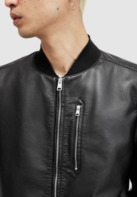 Svart läderbomberjacka med ribbade svarta kragar, med silverdragkedjor fram och en glatt yta. Närbild.