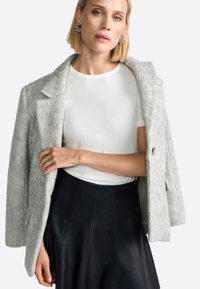 Palton gri din lână cu o textură moale și guler supradimensionat, purtat pe un tricou alb ajustat și o fustă din piele neagră. Detaliu notabil cu nasturi.
