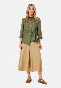 Olivenfarbendes Wickelhemd mit langen Ärmeln, Knopfleiste und Bindedetail, kombiniert mit beigen weiten Hosen und braunen Sandalen.