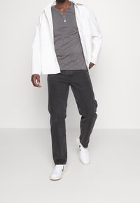 Grijze henley shirt onder een witte lichte jas, gecombineerd met donkere jeans en witte sneakers. Eenvoudige, casual stijl met strakke lijnen.