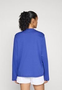 Ilga rankovė mėlynas sportinis marškinėlis su apvalia iškirpte, lygia tekstūra ir plokščiomis siūlėmis, derinamas su baltomis šortais. Paprastas, funkcionalus dizainas.