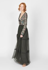 Vestido largo de color verde oscuro con un escote en V profundo, un bodice floral adornado y una falda de capas transparentes con patrones de puntos y detalles en negro.