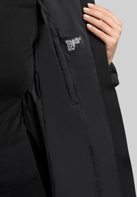 Schwarze gepolsterte Jacke mit glattem Außenmaterial, das ein dezentes Logo-Patch und eine Seitentasche aufweist. Das Innenfutter ist weich und hat ein mattes Finish.