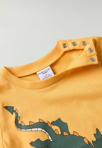 Gul bomulds t-shirt med et grønt dinosaurgrafik. Har trykknapper på skulderen og en rund halsudskæring. Plejeetiket synlig.