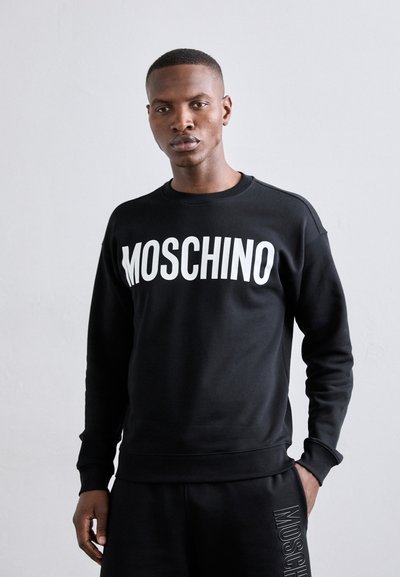 Crni dukserica s podebljanim bijelim logotipom "MOSCHINO" na prednjoj strani, okruglog izreza i rebrastim manžetama i rubom. Izrađena od glatke pamučne tkanine.