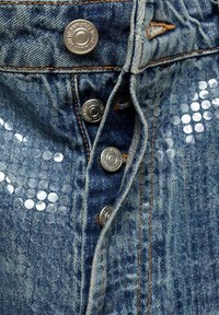 Denim tkanina s teksturiranim uzorkom, koja sadrži pet srebrnih metalnih dugmadi i okrugle šljokice, u srednje plavoj boji.